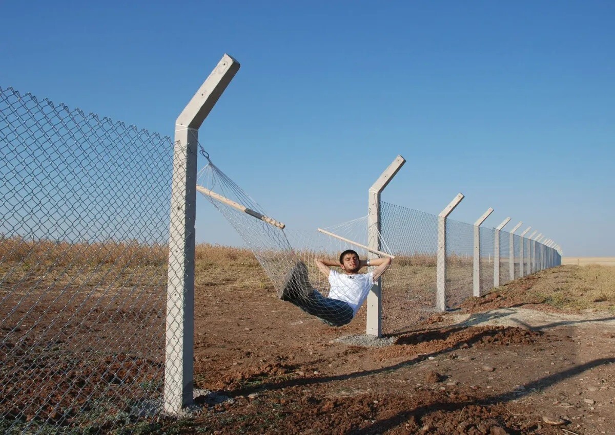 Man lies on a hamack that replaces a piece of fence at the border.