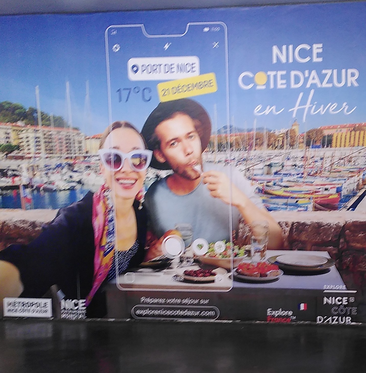 Pub dans le métro ventant "nice côte d'azur en hiver" avec deux personnes en T-shirt en terrasse au bord du port de Nice en train de manger des salades et autres trucs estivaux. Une capture d'écran de portable d'une appli météo indique "port de Nice <br />17°C 21 décembre"